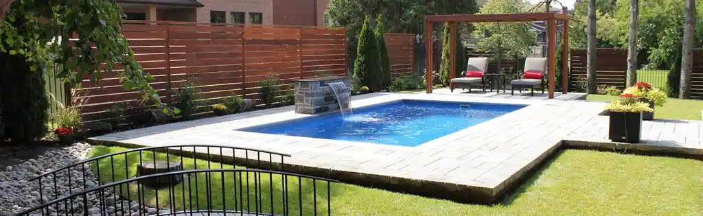 Rectangular fiberglass pool with a stone waterfall feature, paver surround, cedar fence and a timber pergola