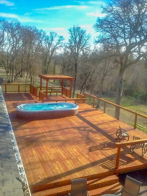 Swim spa set into a wooden deck above a river, with railings and trees behind